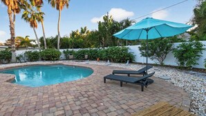 En swimmingpool med liggestole og en blå parasol, omgivet af en murstens terrasse og grøn vegetation.