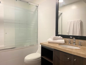 A modern bathroom with a glass shower enclosure, a white toilet, a sink with a beige countertop, and a mirror with a towel hanging on the wall. - Redibuk - Luxury duplex 1BR Miraflores (Lima)