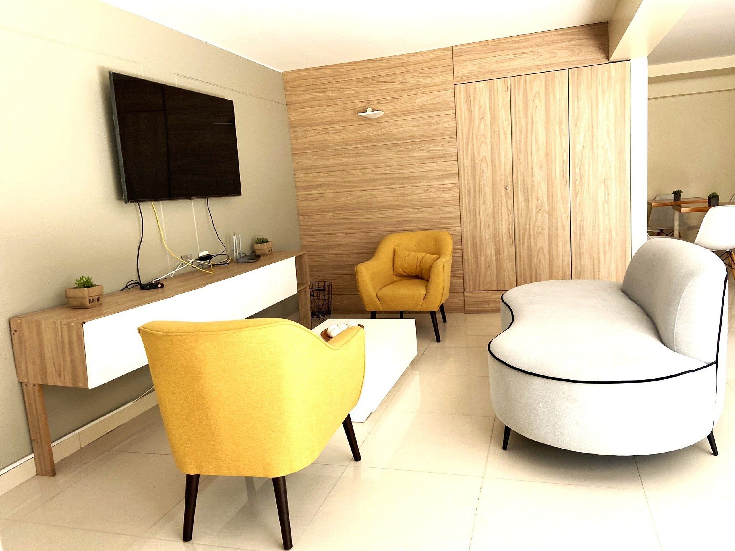 A modern living room with a flat-screen TV mounted on the wall, a wooden console table, a yellow armchair, a white armchair, and a wooden wardrobe.