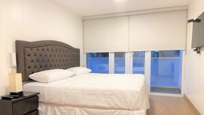 A bedroom with a large bed, a nightstand, a lamp, and a view of the outside through a sliding door. - Redibuk - Luxury duplex 1BR Miraflores (Lima)