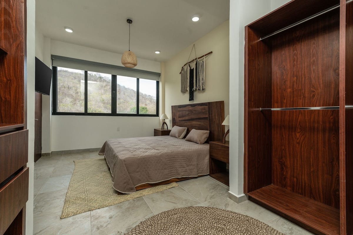 A bedroom with a bed, wooden wardrobe, and a large window.