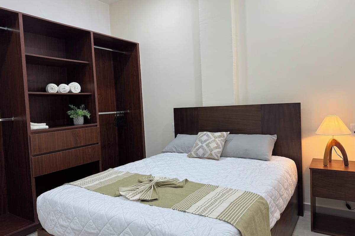 A neatly made bed with a quilted white duvet, a green throw blanket, and a patterned pillow. A wooden wardrobe with multiple shelves is to the left, and a bedside table with a lamp is to the right.