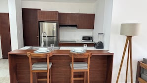 Una cocina moderna con gabinetes de madera, un refrigerador de acero inoxidable, un microondas y una licuadora en la encimera. Hay dos taburetes de bar de madera con asientos acolchados y una pequeña isla con un lavamanos y dos vasos.