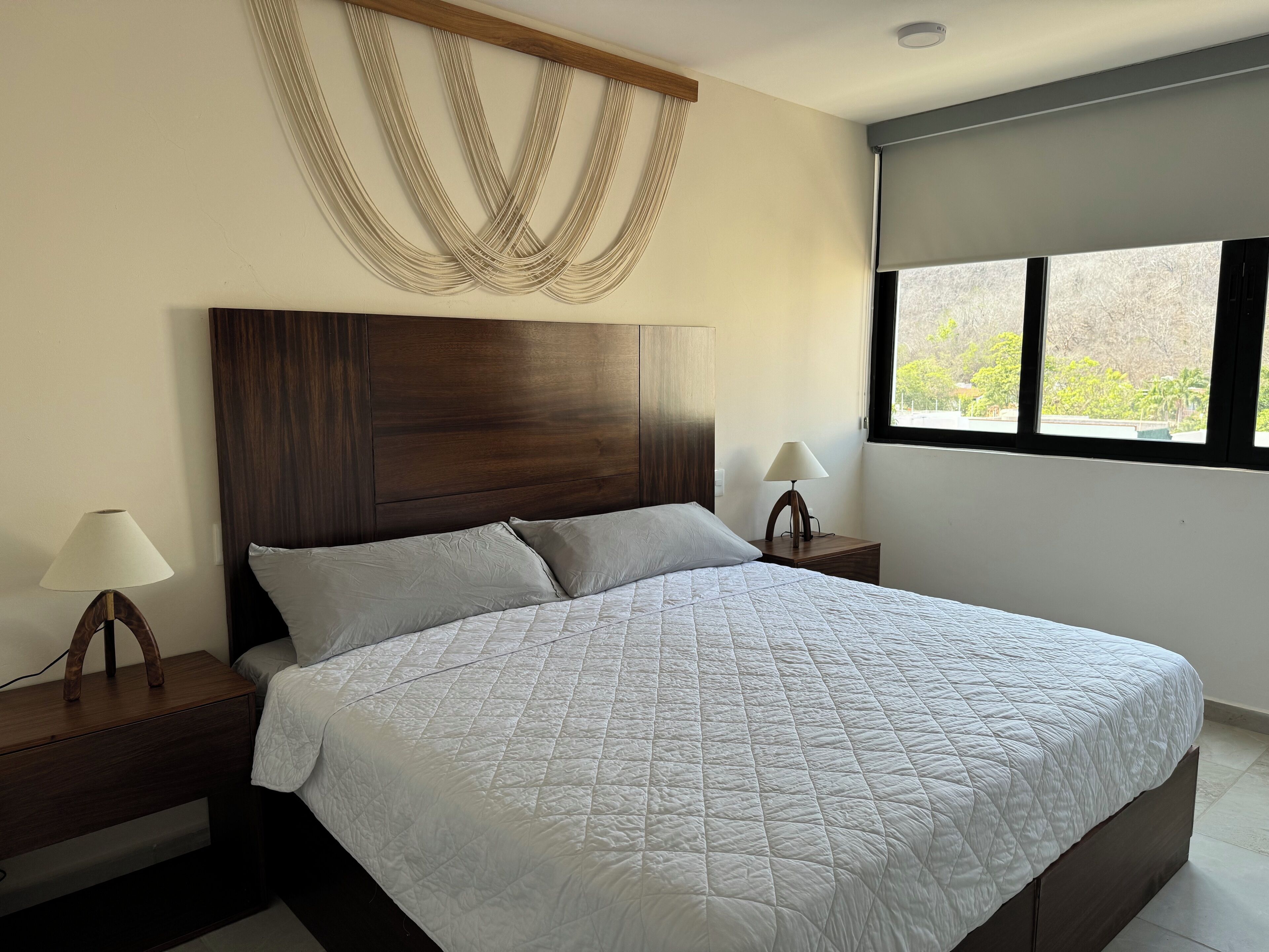 Une chambre à coucher avec un grand lit, une tête de lit en bois, deux tables de chevet avec des lampes et une fenêtre donnant sur un paysage verdoyant.