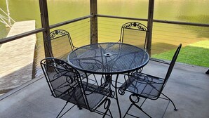 A covered outdoor patio with a round metal table and four chairs, overlooking a body of water and a forested area.