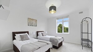 A bedroom with two beds, a window, a hanging light fixture, and a wall-mounted shelf.