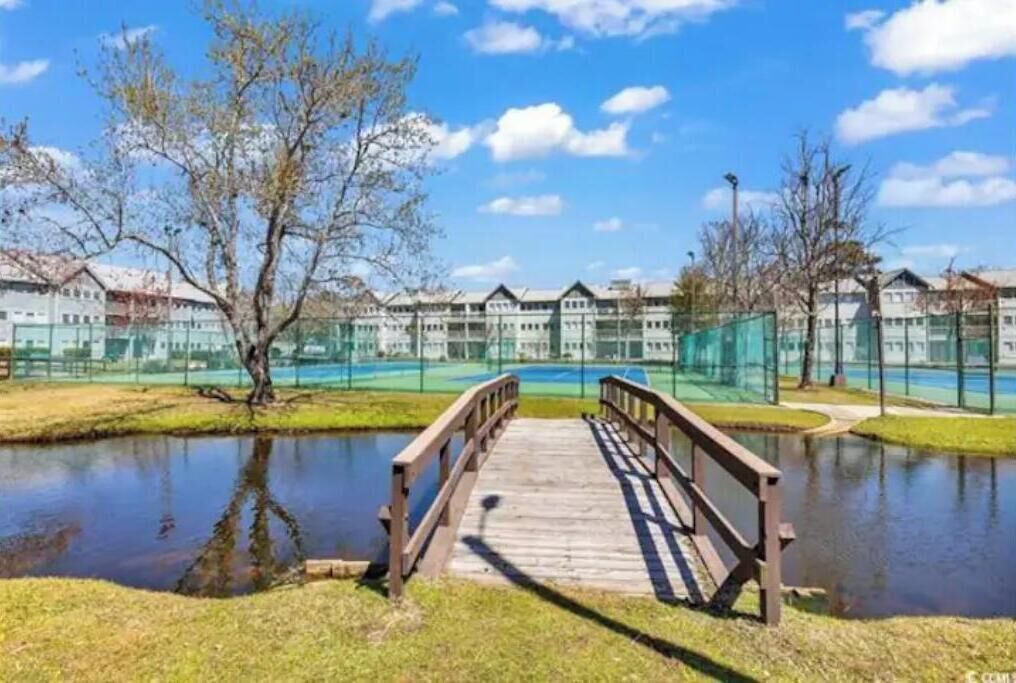 A wooden bridge over a calm pond, leading to a row of buildings with a clear blue sky above.