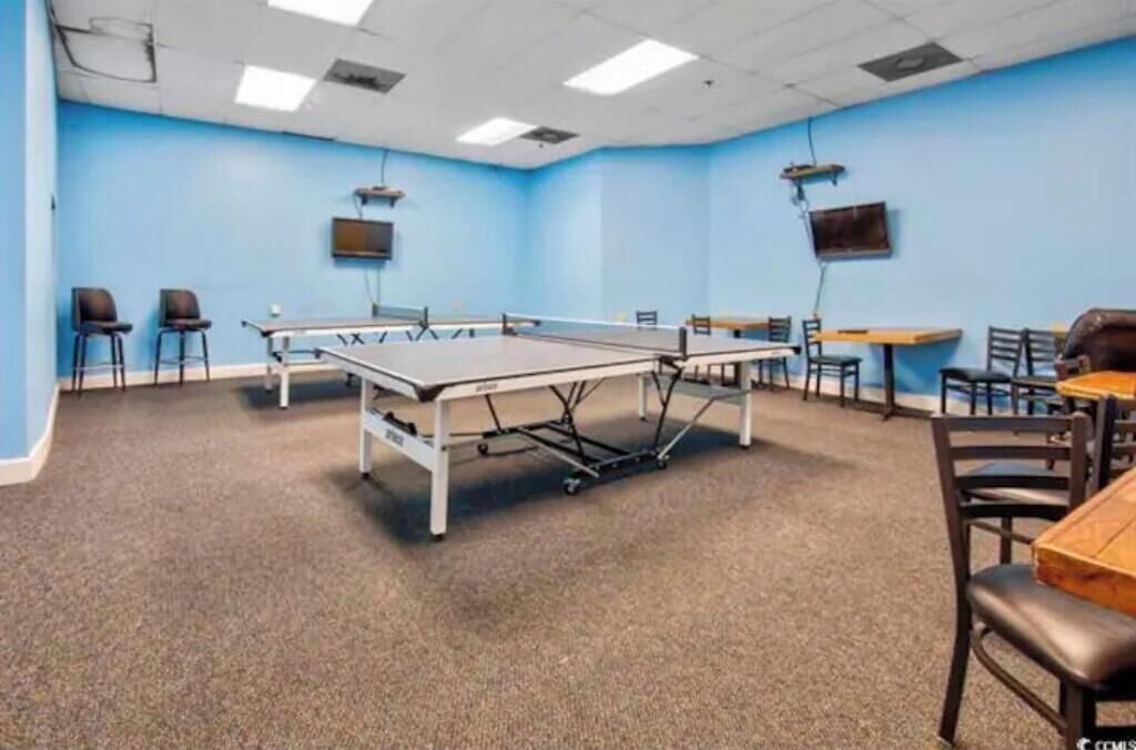 A room with a ping pong table, chairs, and tables, with blue walls and carpeted flooring.