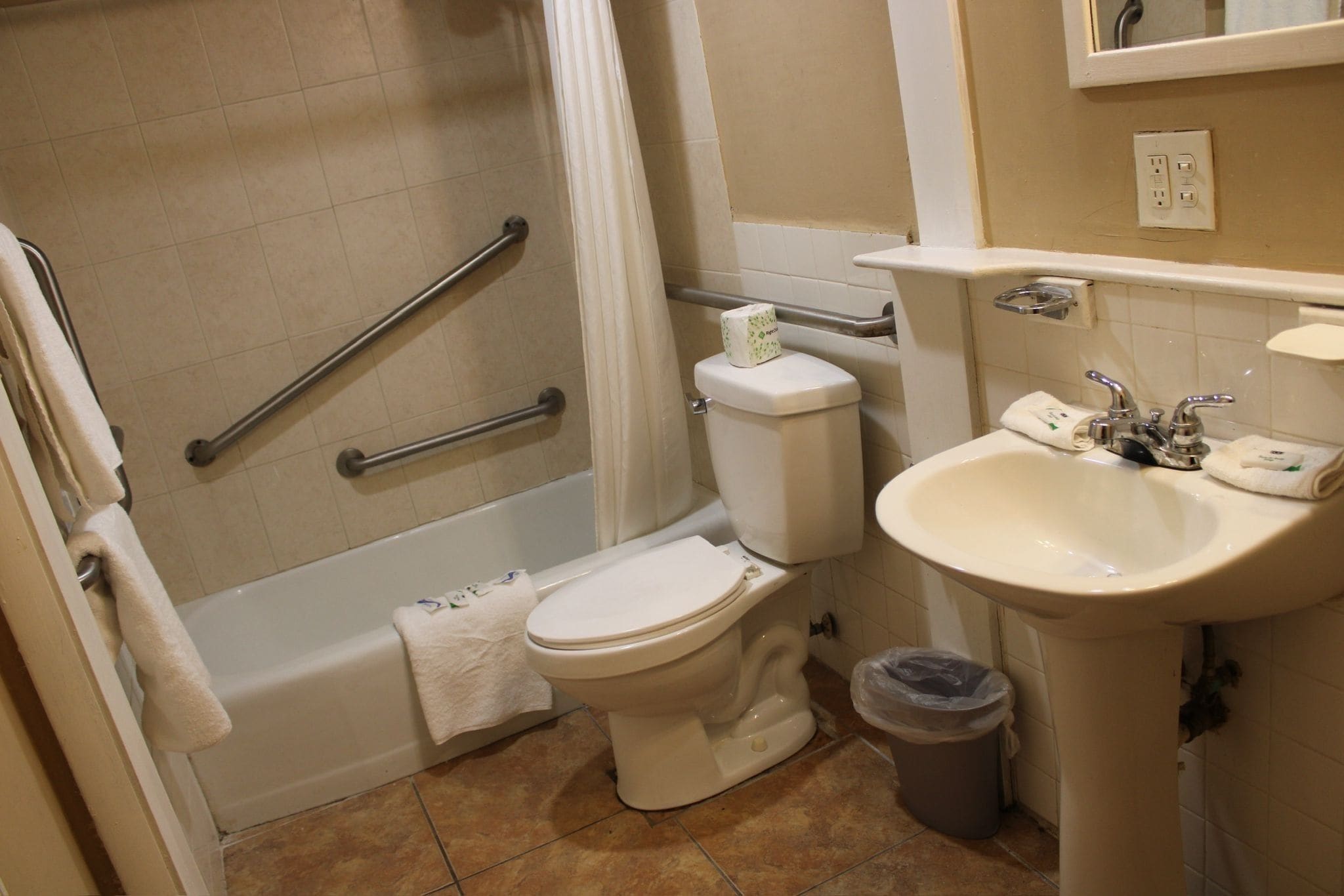 A bathroom with a toilet, sink, and bathtub. There are grab bars next to the bathtub and a trash can near the sink.