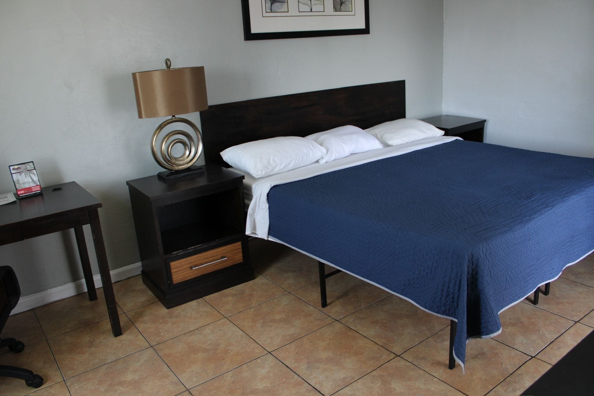 A neatly made bed with a blue bedspread, a dark wooden headboard, a bedside lamp, and a small drawer unit.