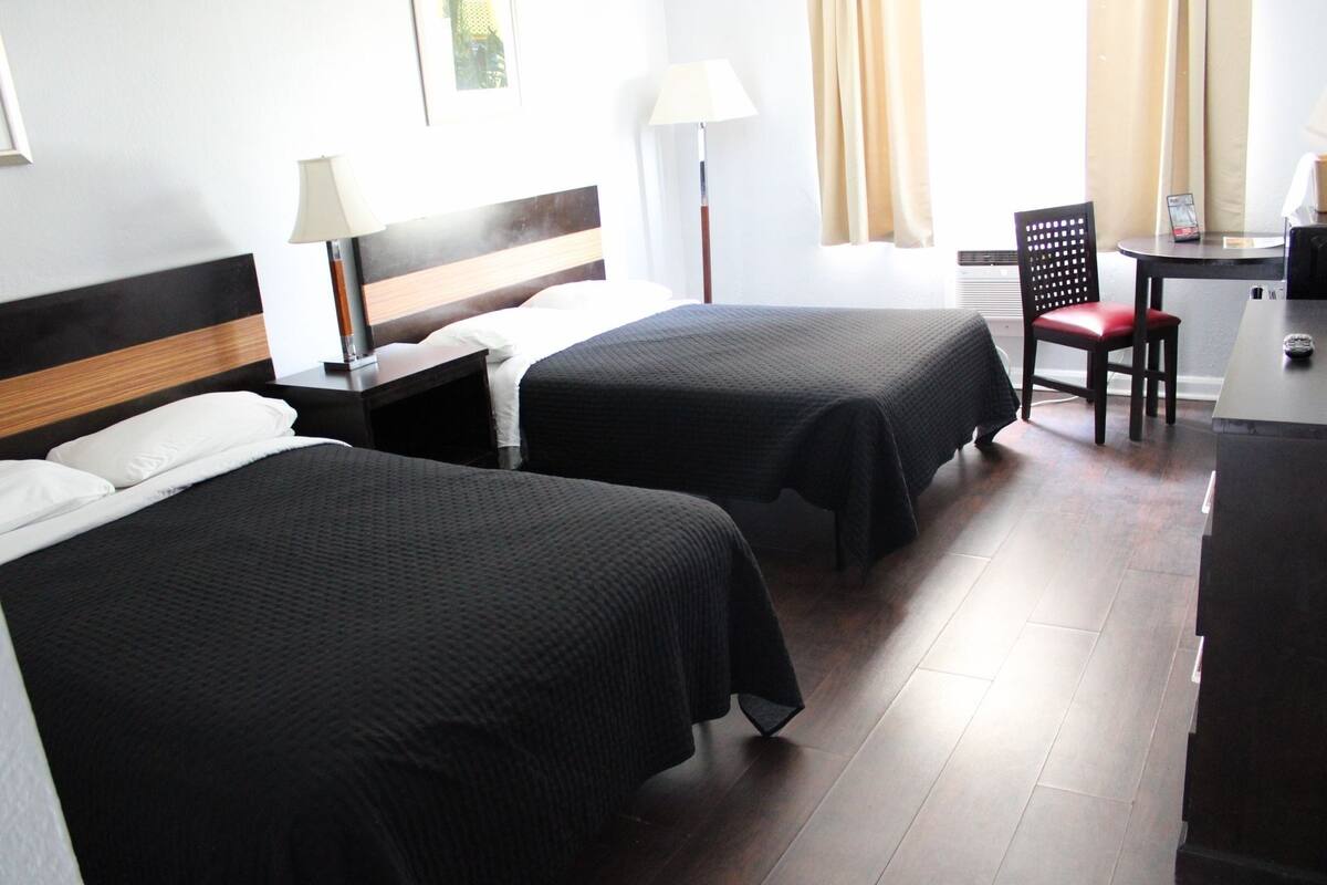 A hotel room with two beds, a desk, a chair, and a lamp.