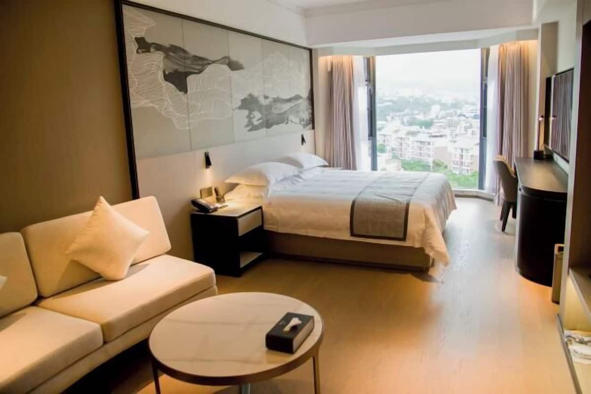 A hotel room with a bed, a sofa, a small round table, a TV, and a large window with a city view.