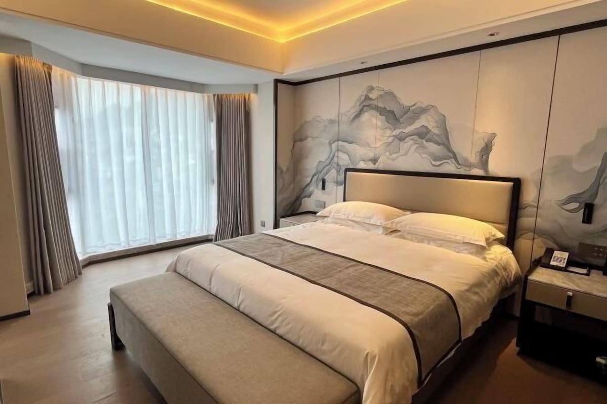 A modern hotel room with a large bed, a bench, a nightstand, and a wall with a mountain painting.