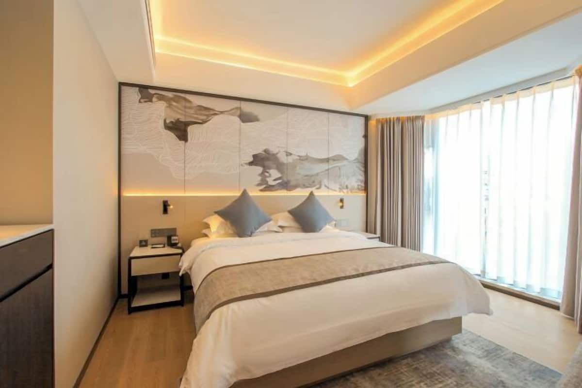 A modern hotel room with a large bed, a nightstand, a wall-mounted artwork, and a large window with curtains.