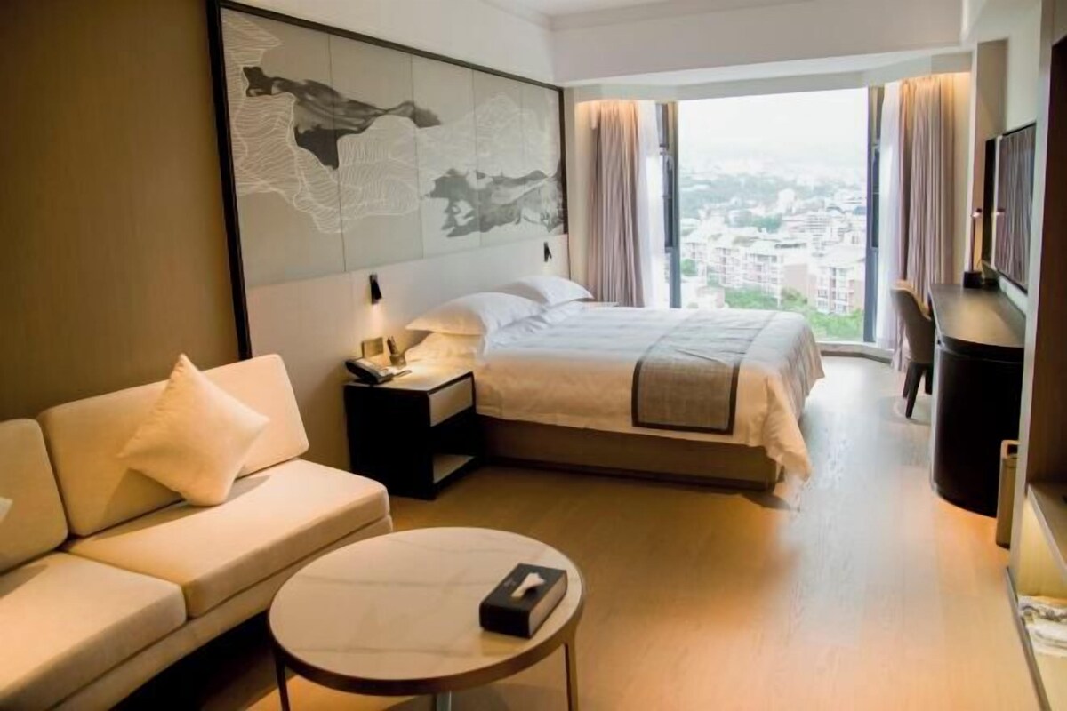 A hotel room with a bed, a sofa, a bedside table, a small round table, and a large window offering a city view.