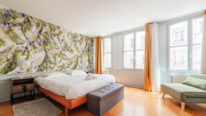 A modern bedroom with a wooden floor, a bed with a grey bench, a nightstand, a large window with curtains, and a colorful abstract wall. - La Place de la Cathédrale - Superbe appt face à la Cathédrale (Strasbourg)