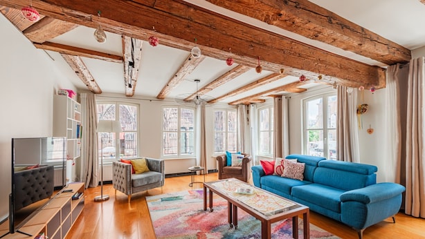A spacious living room with a blue sofa, a grey armchair, and a wooden coffee table. The room has a wooden ceiling with exposed beams and large windows with curtains. - La Place de la Cathédrale - Superbe appt face à la Cathédrale (Strasbourg)