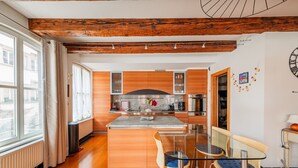 A modern kitchen with wooden cabinets, a central island, and a dining area with a glass table and chairs. - La Place de la Cathédrale - Superbe appt face à la Cathédrale (Strasbourg)