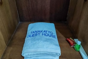 A folded towel with 'TANAKATTO GUEST HOUSE' text, a red packet, and a green bottle on a wooden surface. - RedDoorz @ Tanakatto Homestay Waikabubak (Waikabubak City)