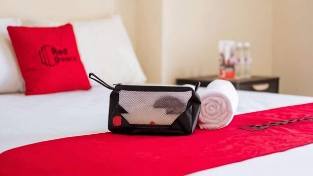 Habitación de hotel con cama, una manta roja, una toalla enrollada, almohada roja y un objeto negro pequeño sobre la cama.