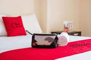 Habitación de hotel con cama, una manta roja, una toalla enrollada, almohada roja y un objeto negro pequeño sobre la cama.