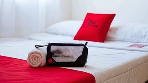 Una cama bien hecha con una almohada roja, una toalla enrollada y una bolsa de malla negra sobre la cama.
