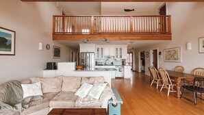 Een ruime woonkamer met een hoge plafond, een houten balkon, een keuken met witte kasten, en een eethoek met houten stoelen.