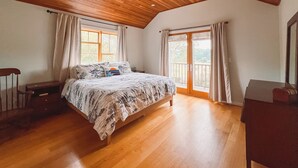 Een slaapkamer met een houten plafond, een bed met een bloemenbedekking, twee ramen met gordijnen en een openslaande deur naar een balkon.