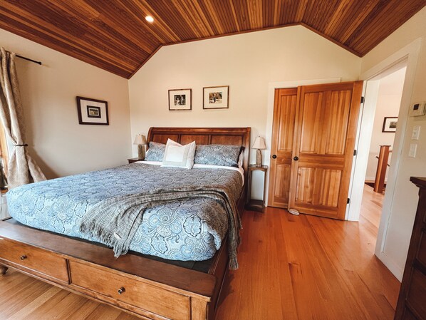 A bedroom with a wooden ceiling, a large bed with patterned bedding, two bedside tables with lamps, and a wooden door.