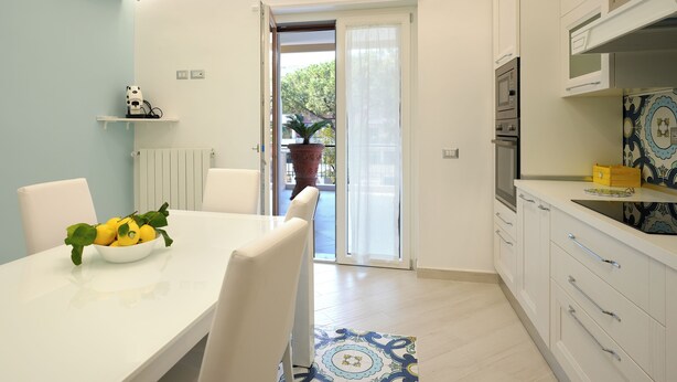 Uma cozinha moderna com armários brancos, forno embutido e micro-ondas. Há um espaço de refeições com mesa e cadeiras brancas, e uma tigela de frutas sobre a mesa.
