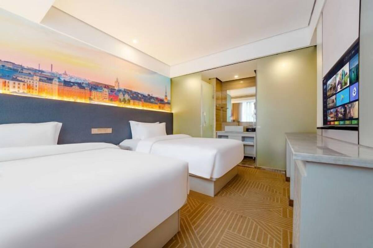 A hotel room with two beds, a cityscape mural, a desk, and a TV.