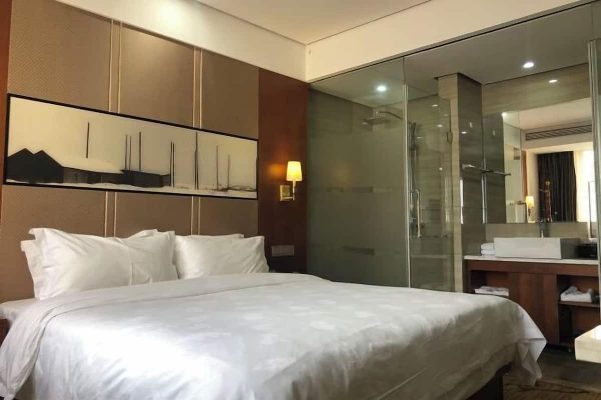 A hotel room with a large bed, a glass shower enclosure, a wooden cabinet, and a wall-mounted mirror.