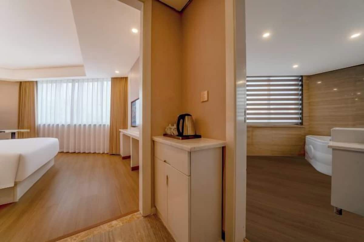 A hotel room with a bed, a small kitchenette, a bathroom, and a window with blinds.
