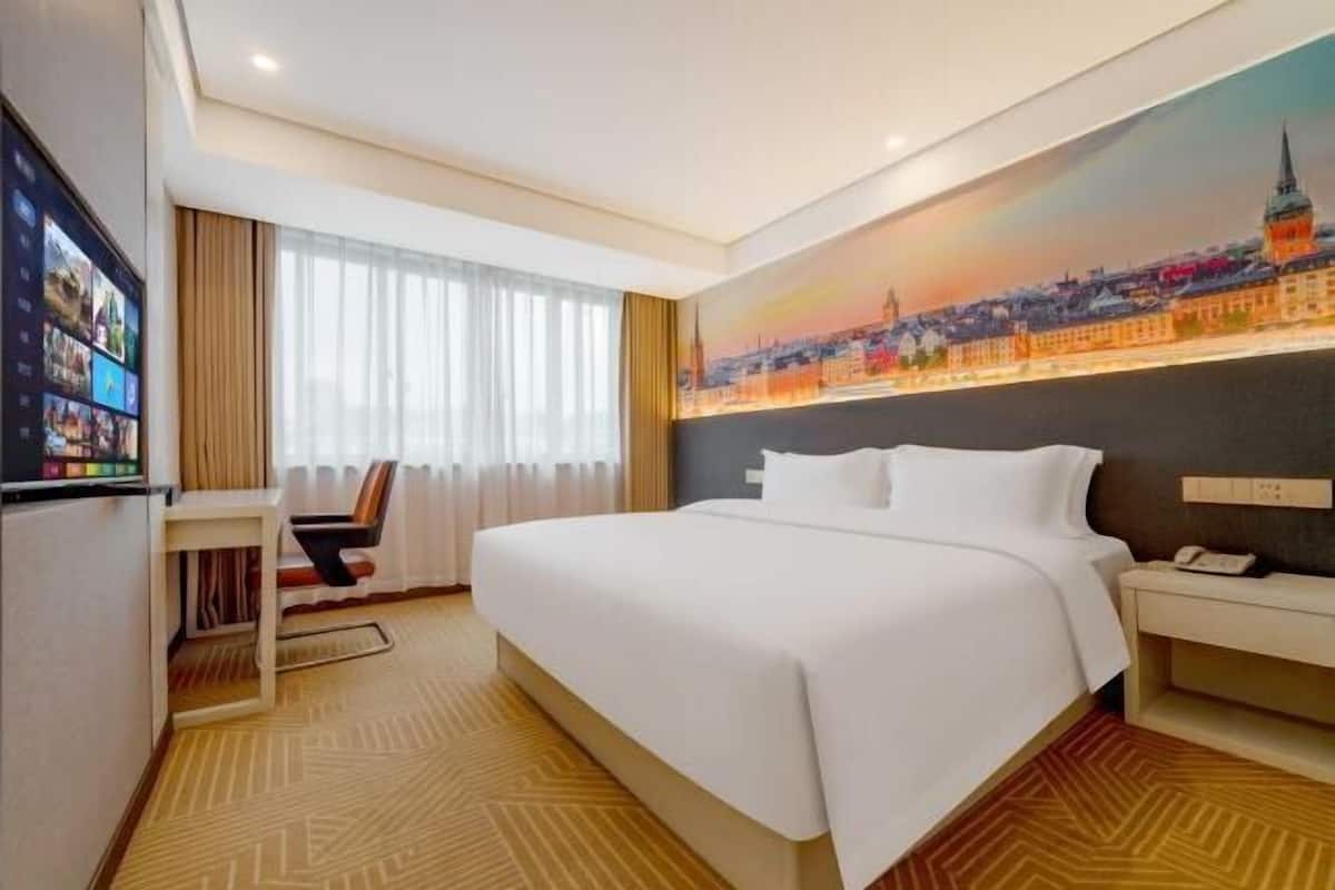 A hotel room with a large bed, a desk, a chair, a TV, and a cityscape painting on the wall.