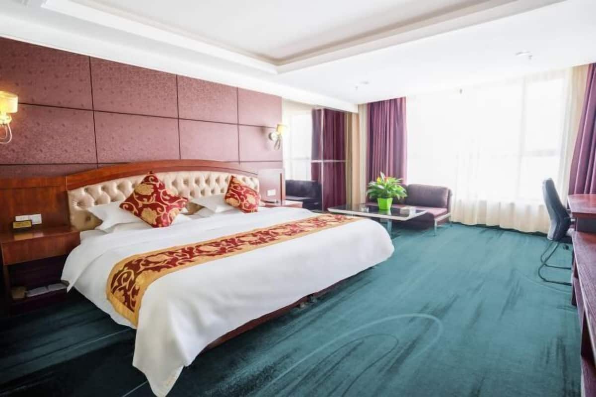 Habitación de hotel con una cama grande, cabecera de madera, alfombra verde, escritorio con una silla y una ventana con cortinas.