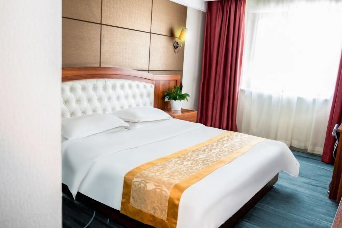 A hotel room with a large bed, a wooden headboard, a bedside table with a lamp, and a window with curtains.
