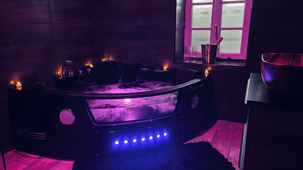 Une pièce faiblement éclairée, avec un grand jacuzzi circulaire au centre, entouré de bougies et agrémentée d’une fenêtre à verre dépoli.