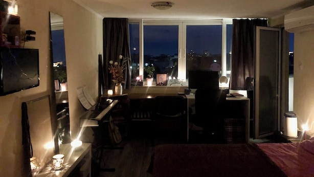 Et hotelværelse med skrivebord, stol og seng. Der er et vindue med gardiner, et vægmonteret fladskærms-tv og et lille bord med et lys.