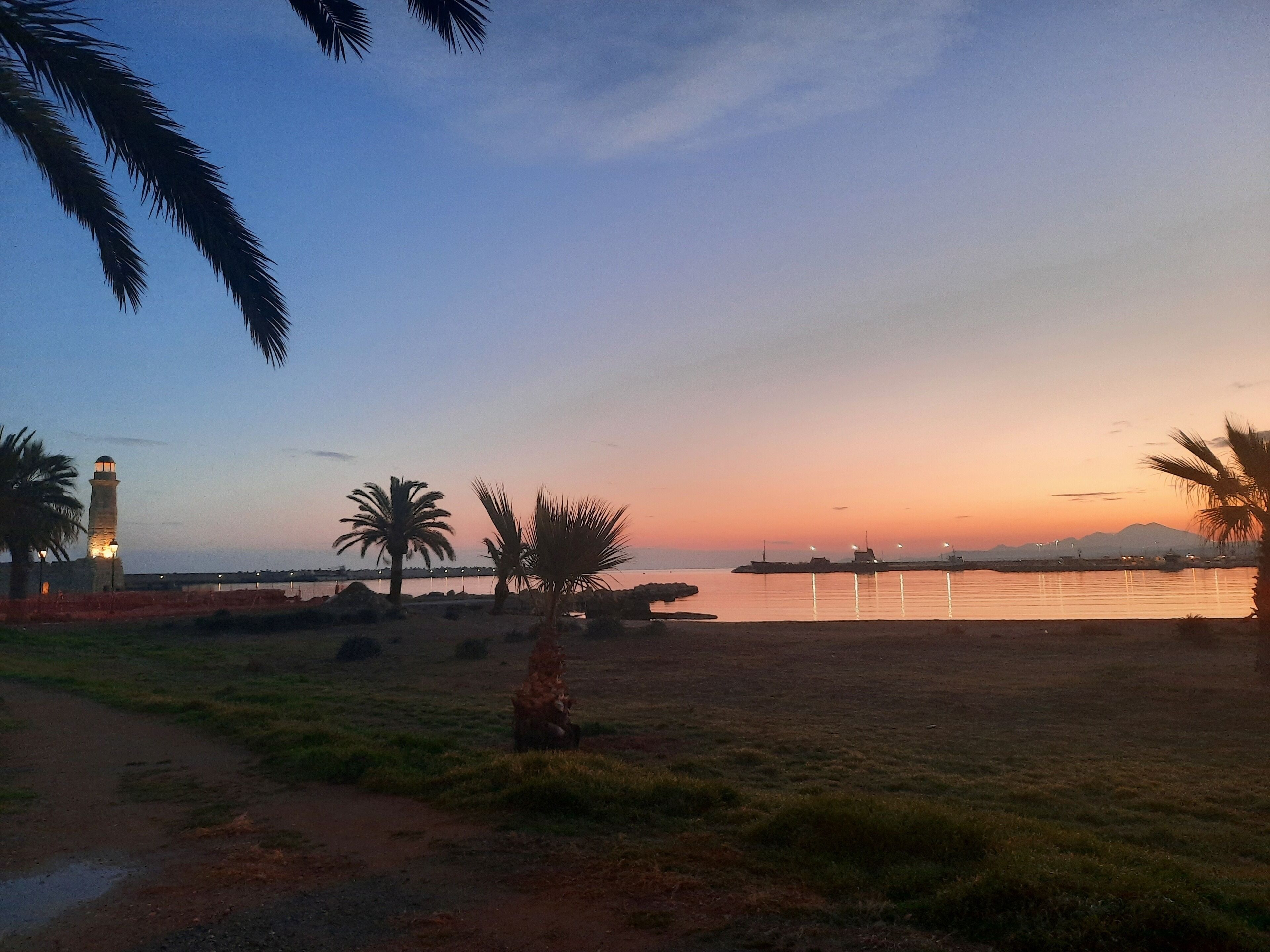 A coastal landscape with a lighthouse, palm trees, and a calm sea at sunset.