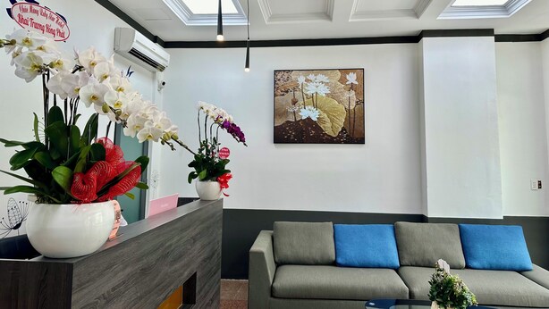 A modern hotel reception area with a wooden reception desk, a grey sofa with blue cushions, and a ceiling fan. - Ruby Star Hotel Nam Quoc Cang (Ho Chi Minh City)