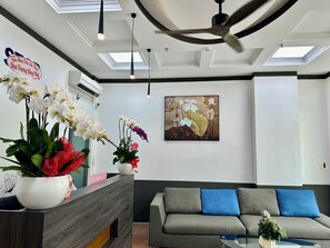 A modern hotel reception area with a wooden reception desk, a grey sofa with blue cushions, and a ceiling fan.