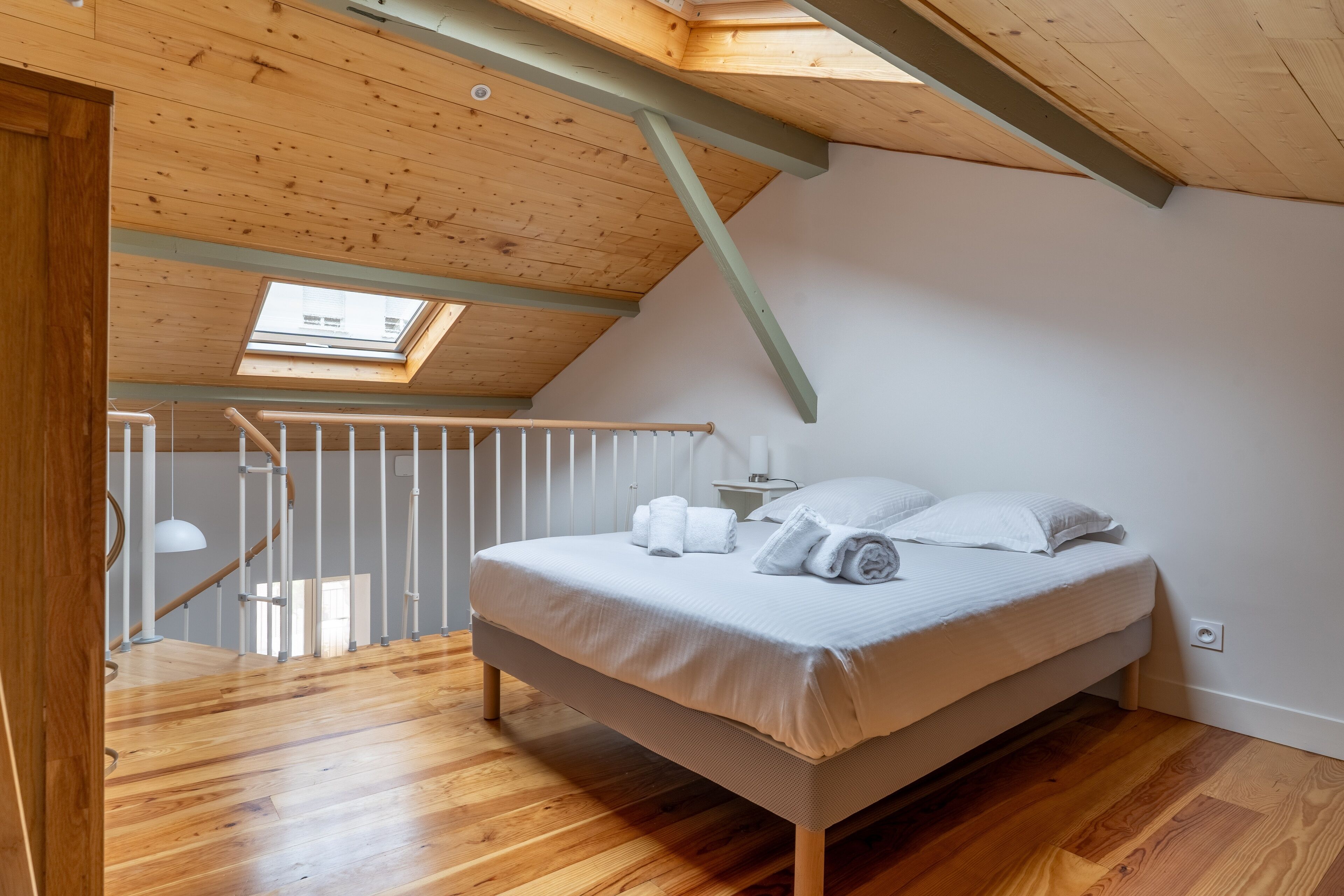 Un dormitorio con techo de madera, tragaluz, cama con ropa blanca y piso de madera.