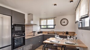A modern kitchen with a built-in oven, a dining table with chairs, and a clock on the wall. - The Blue Rocks - Maison à moins d'1 km de la plage (Lamballe)