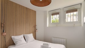 A neatly made bed with white linens, a wooden slat wall, and a window with blinds.
