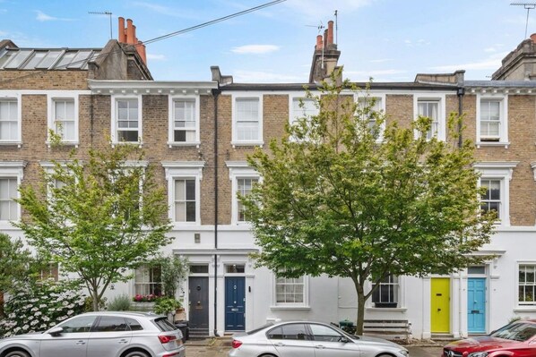 A row of terraced houses with white front doors, some with plants in front, and cars parked in front.