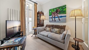 Un salon comprenant un canapé, un téléviseur, une lampe, une bibliothèque et un tableau représentant un chien.