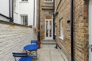 A narrow alleyway with a brick wall, a white door, and a small outdoor seating area with blue chairs and a table.