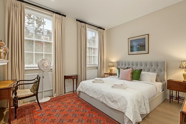 A hotel room with a large bed, a chair, a desk, a radiator, and a window with curtains.