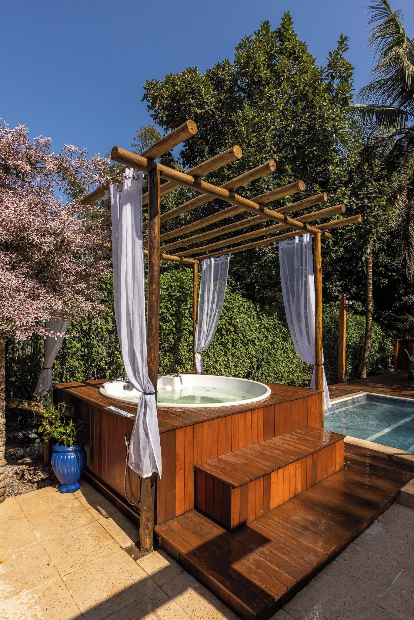 Ein hölzernes Whirlpool mit Pergola-Überdachung, umgeben von üppiger Vegetation und einem Schwimmbad.