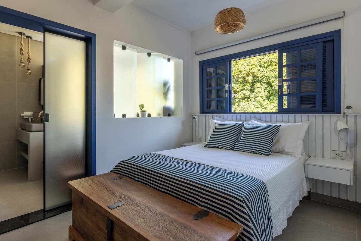 A bedroom with a bed, a wooden bench, a window with blue shutters, and a white bedside table.